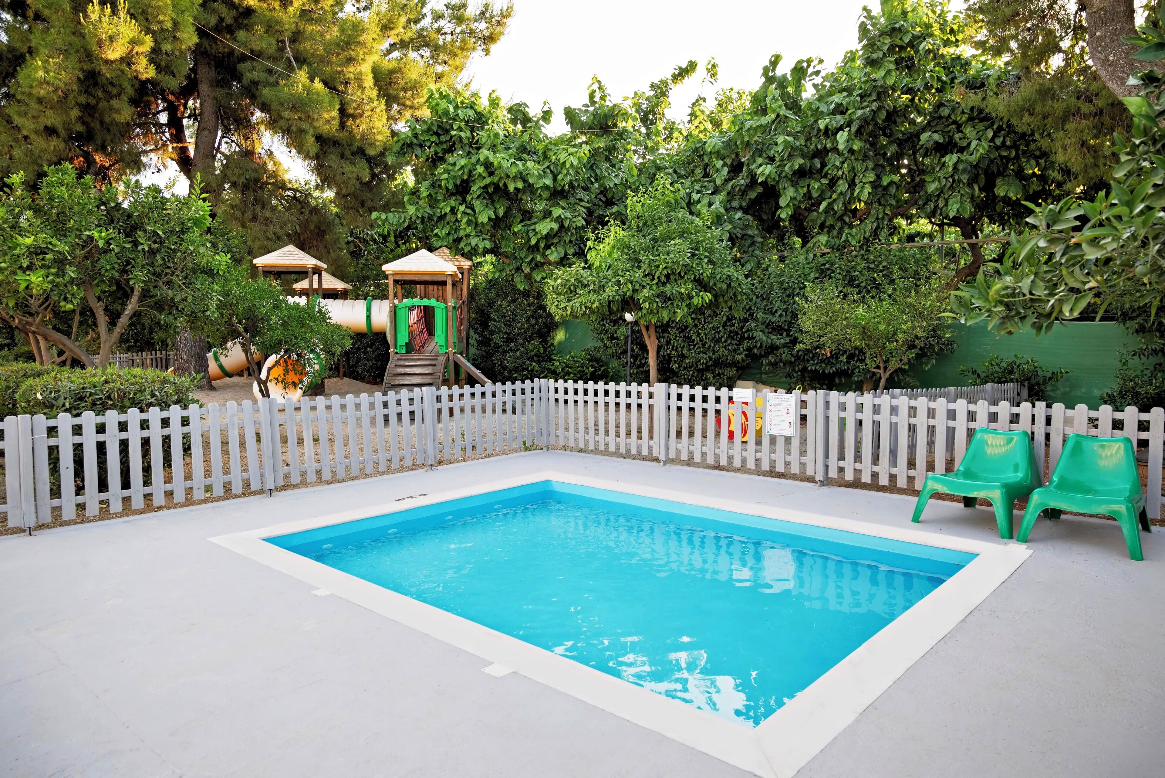Una piscina rectangular con un borde blanco, rodeada por una cerca de piquetes blancos y sillas verdes cerca de la cerca.