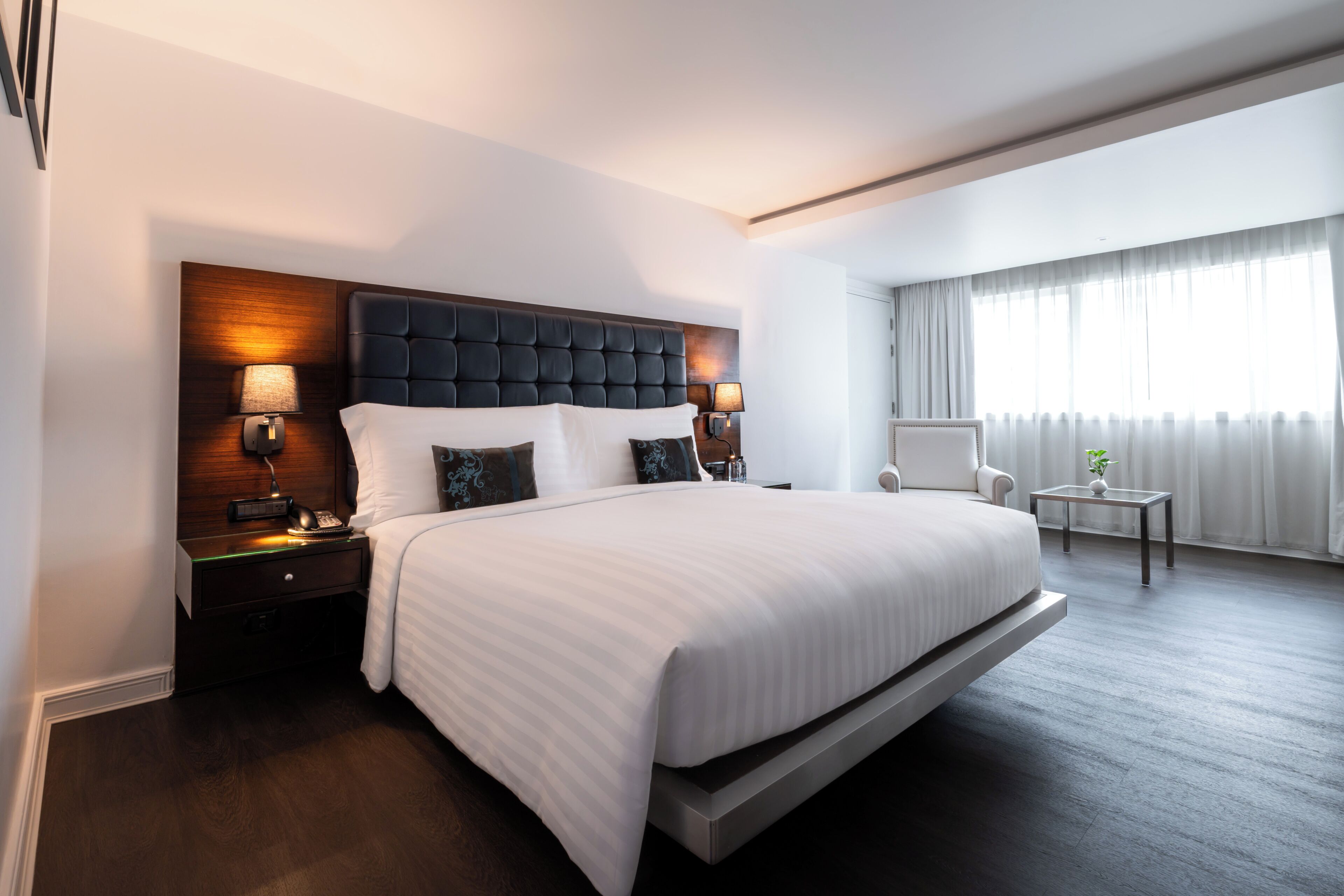 Quarto de hotel moderno com uma cama grande, abajures, uma mesinha e uma cadeira.