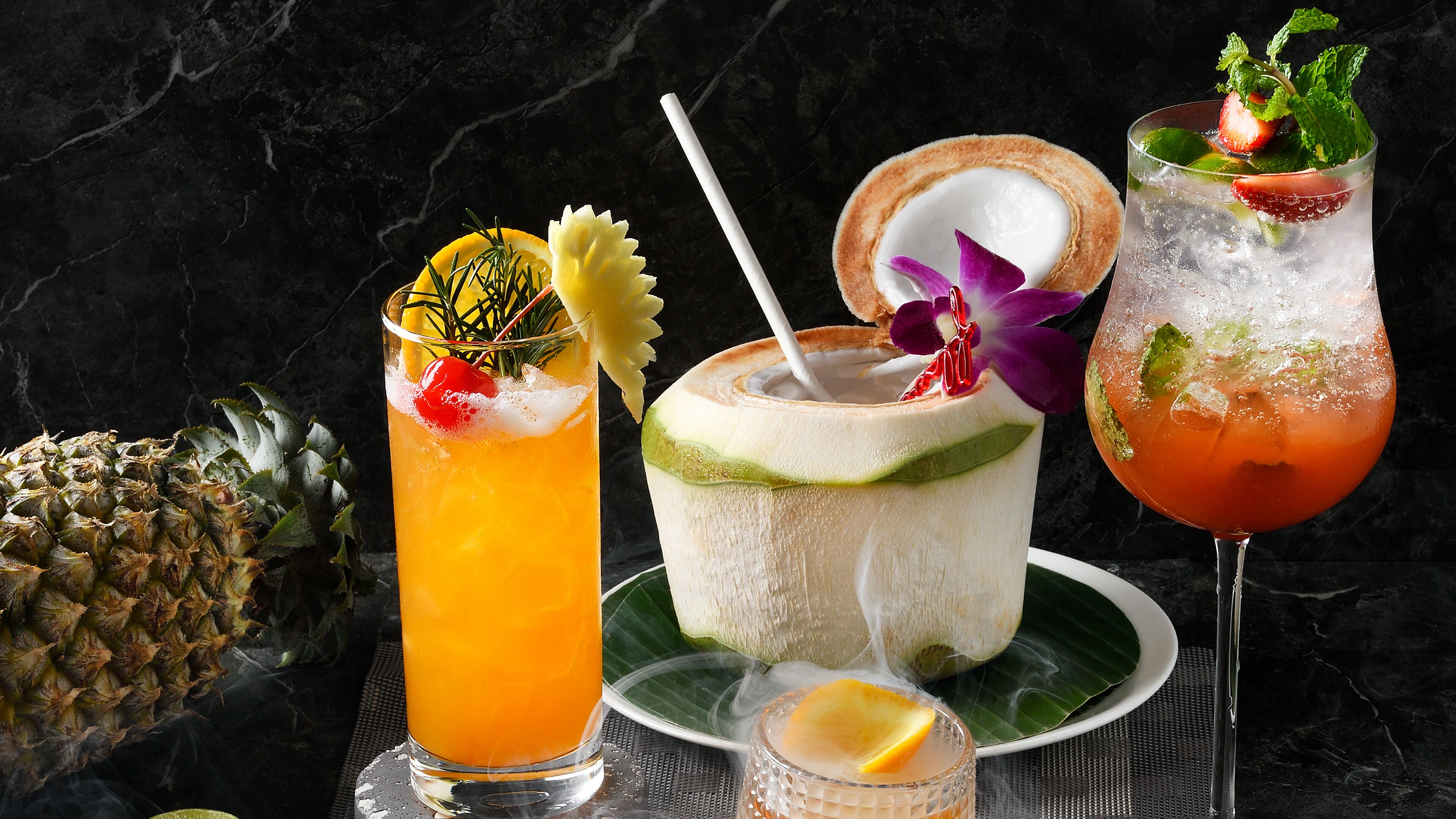 Three drinks with tropical garnishes, a pineapple, and a coconut drink on a dark marble surface.