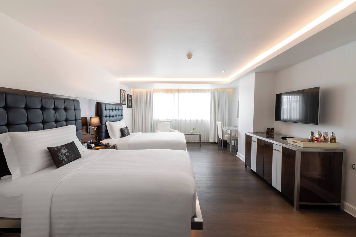 A modern hotel room with two beds, a desk, a flat-screen TV, and a minibar.
