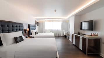 A modern hotel room with two beds, a desk, a flat-screen TV, and a minibar.