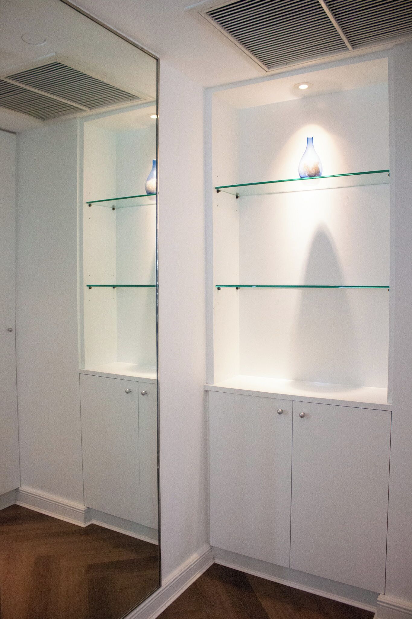 A room with a mirrored wall, a white cabinet with glass shelves, and a wooden floor.