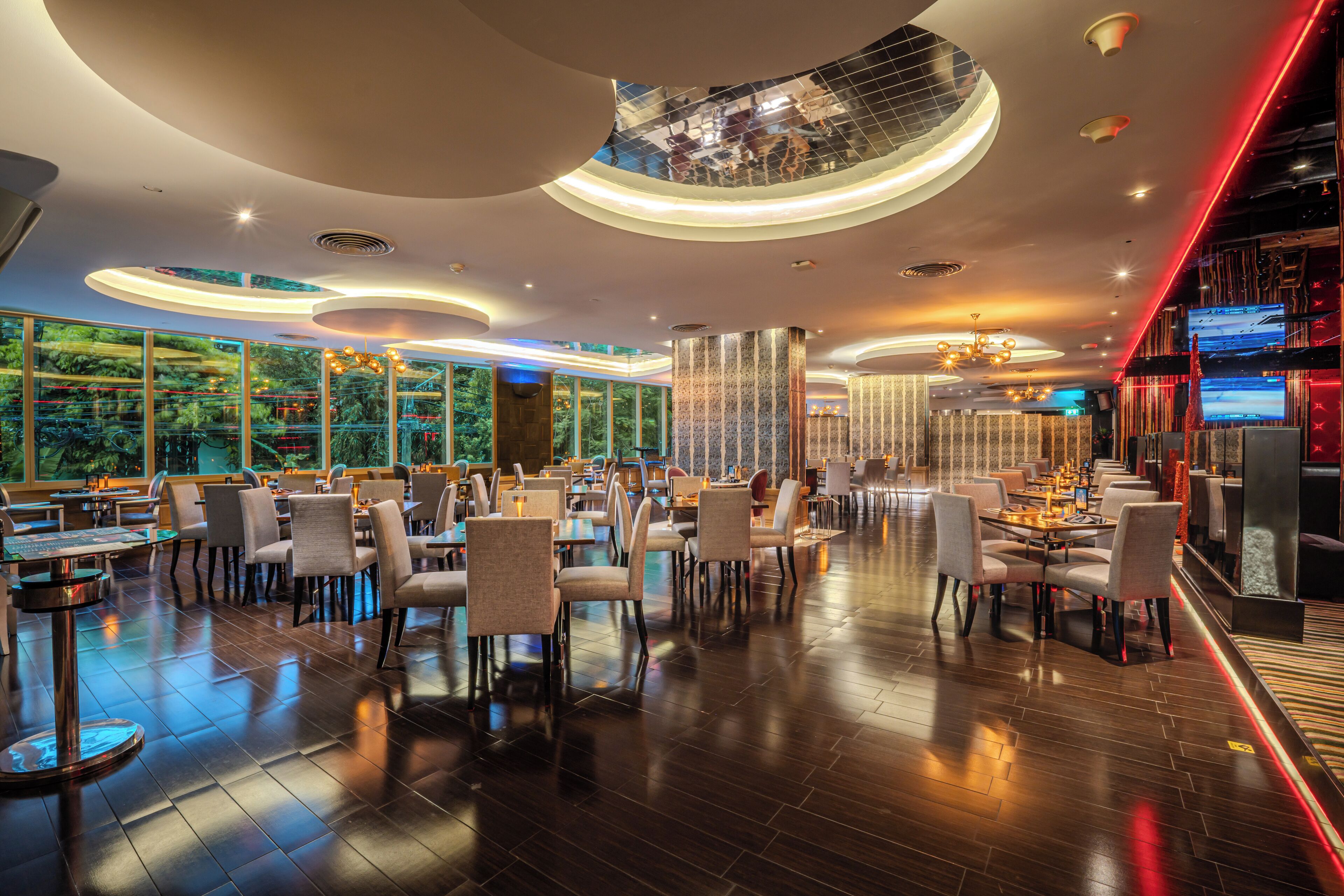 A modern restaurant with a high ceiling, glass walls, and a view of greenery outside.