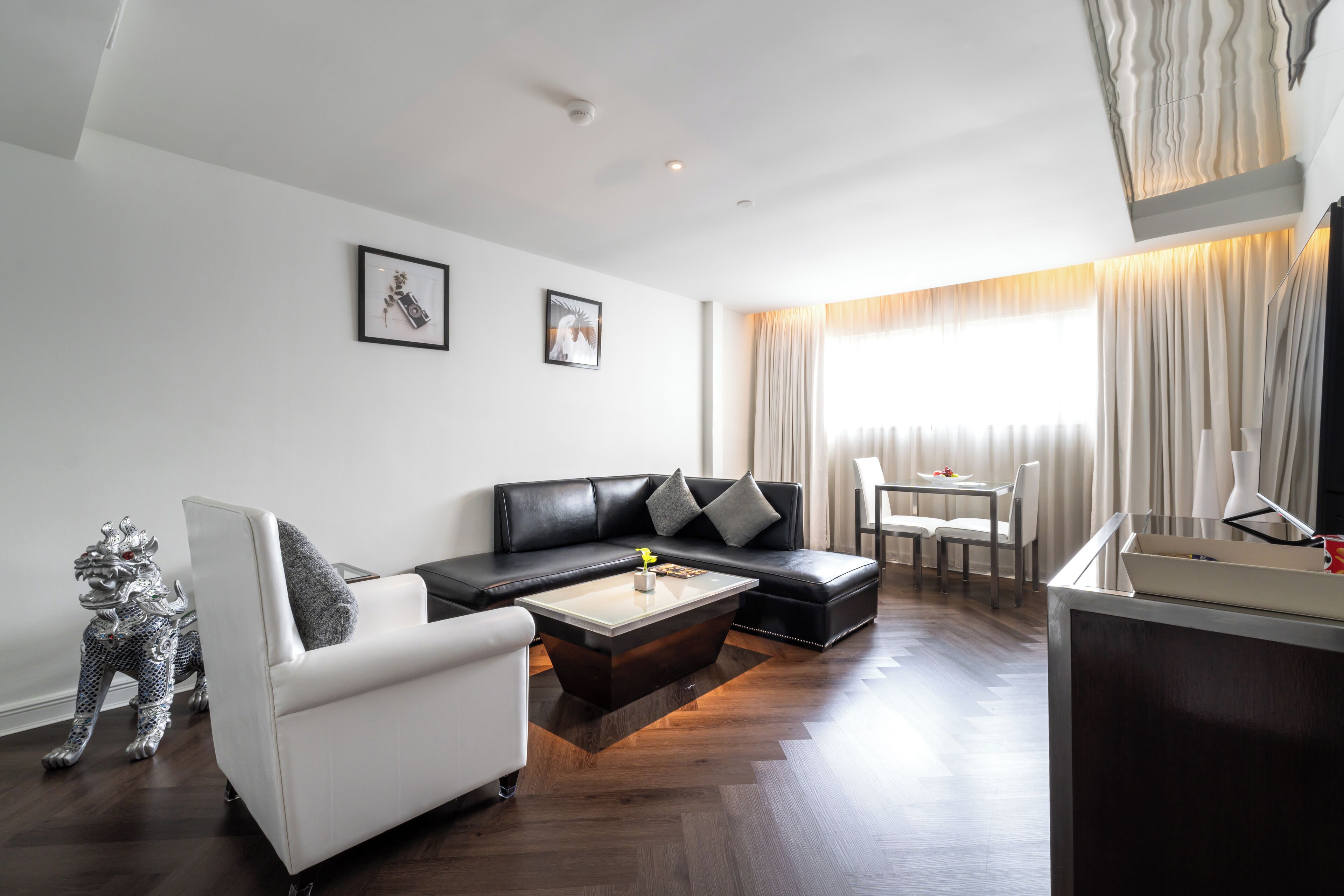 A modern living room with a black leather sofa, white armchair, and a coffee table. There is a small table with a vase and a picture frame. A decorative silver statue is visible on the floor.
