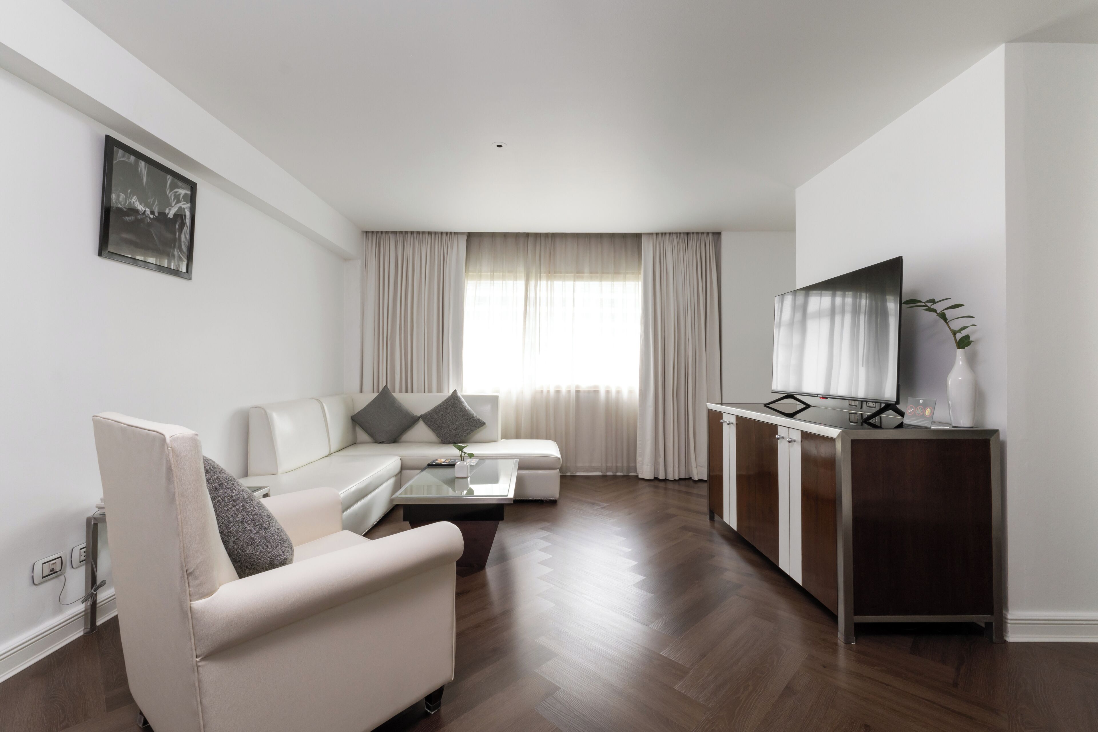 Un salon moderne avec un canapé, un fauteuil et une table basse. On y trouve un téléviseur posé sur un meuble et une fenêtre avec des rideaux.