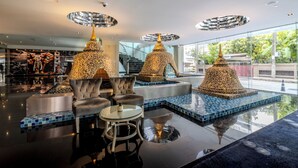 Un vestíbulo moderno con techo reflectante, una zona de estar central con una mesa de vidrio y una fuente decorativa.