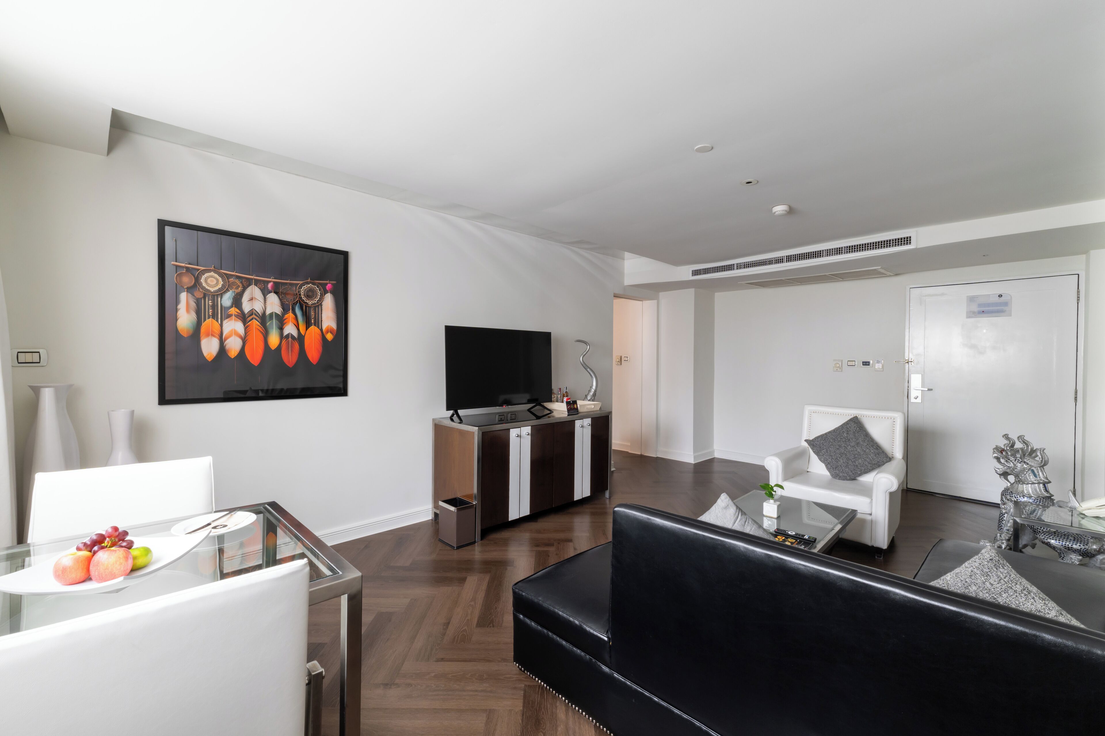A modern living room with a glass dining table, white chairs, a black leather sofa, a flat-screen TV, a cabinet, and a decorative wall piece.