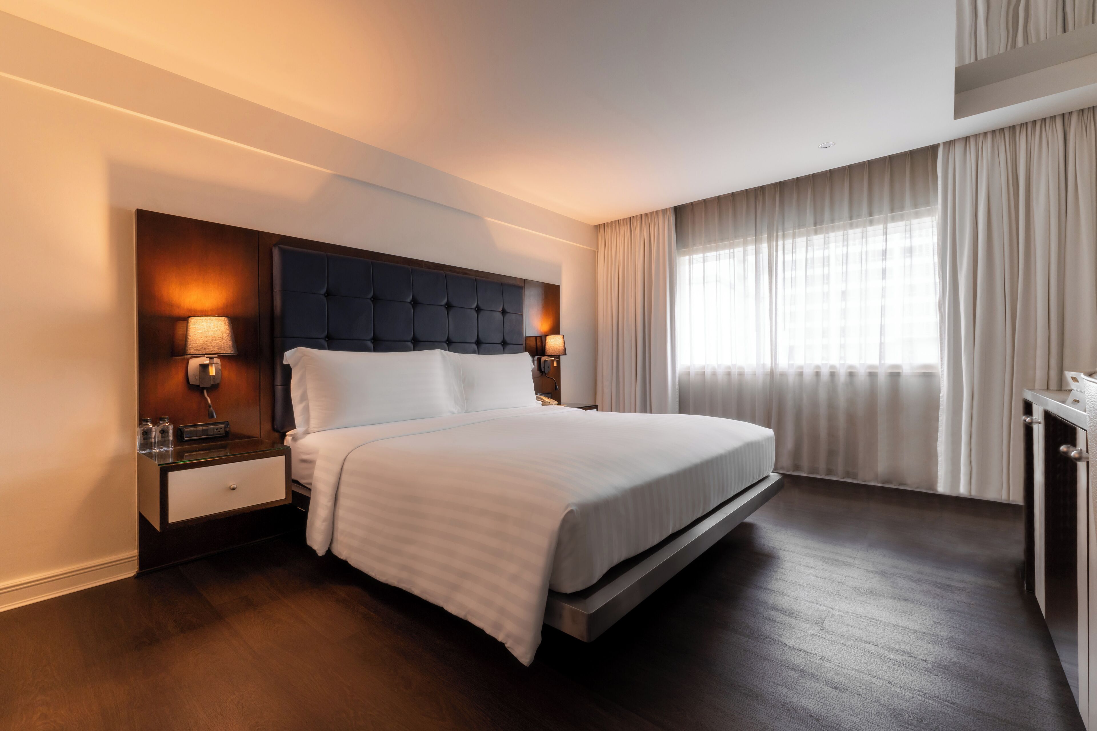 A modern hotel room with a large bed, a nightstand with a lamp, and a view of the city through large windows.