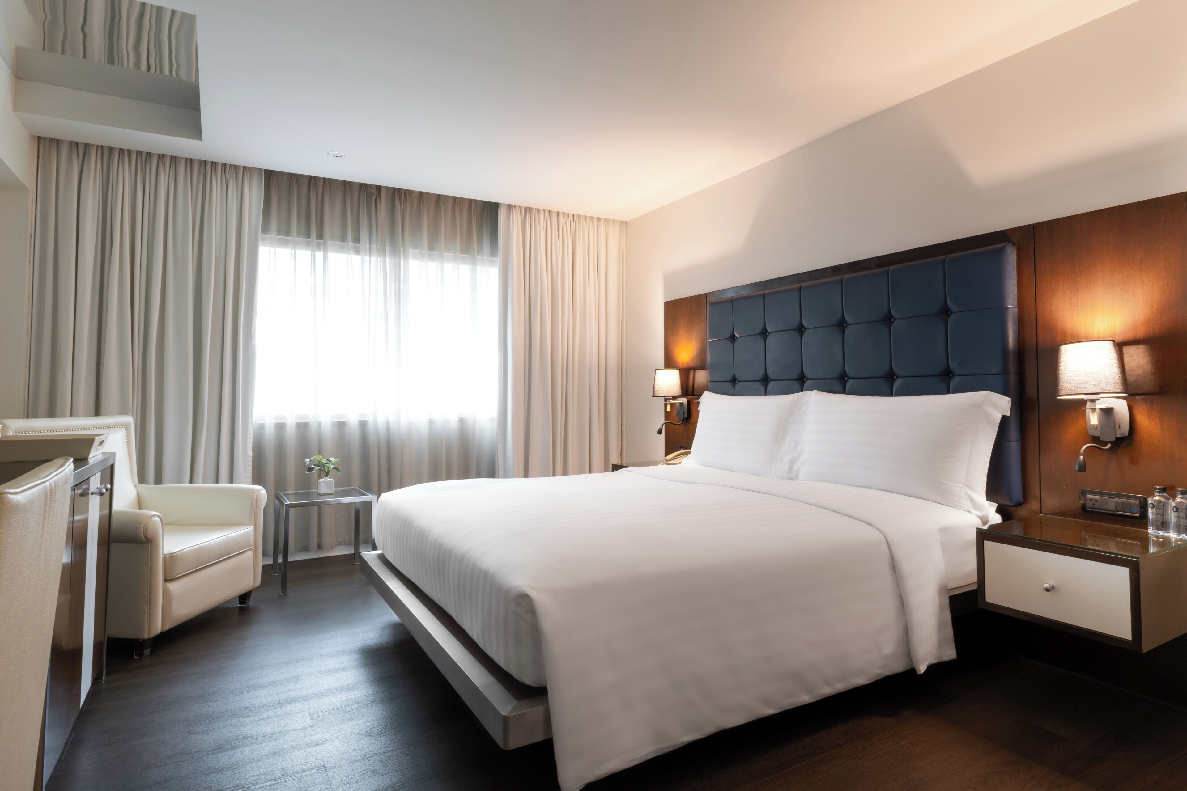Quarto de hotel com uma cama grande, mesas de cabeceira, uma escrivaninha, uma cadeira e uma janela com cortinas.