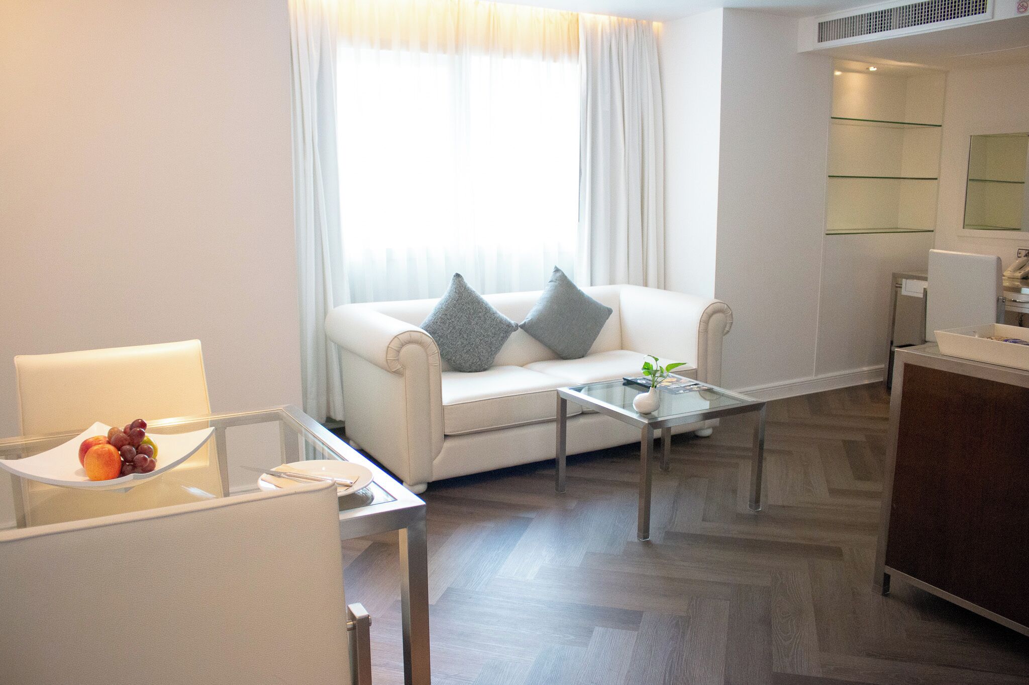 Una sala moderna con un sofá blanco, una mesa de centro de cristal y un comedor con una silla blanca y un plato de fruta.