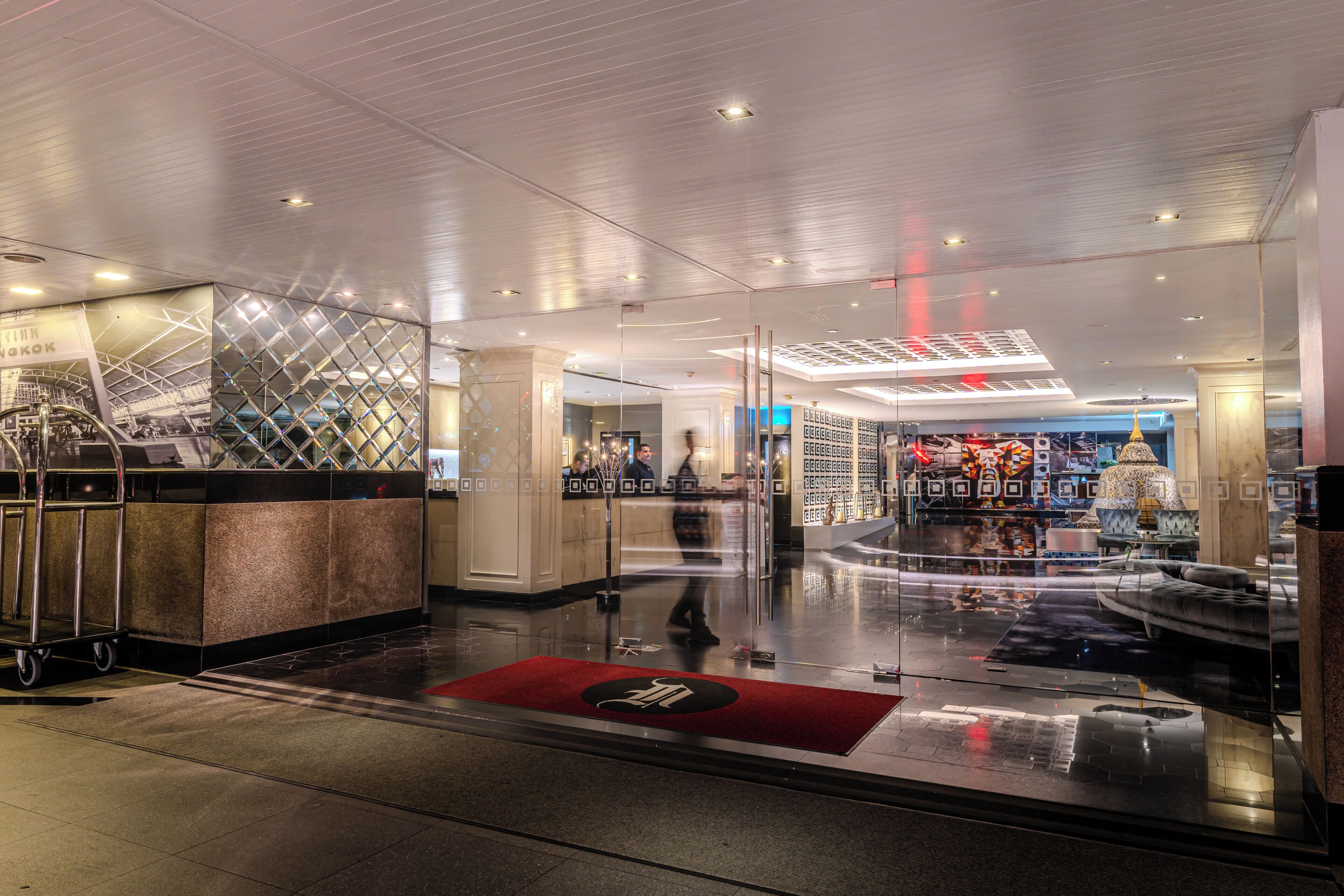 En moderne hotellobby med reception, glasafskærmning og rødt tæppe ved indgangen.