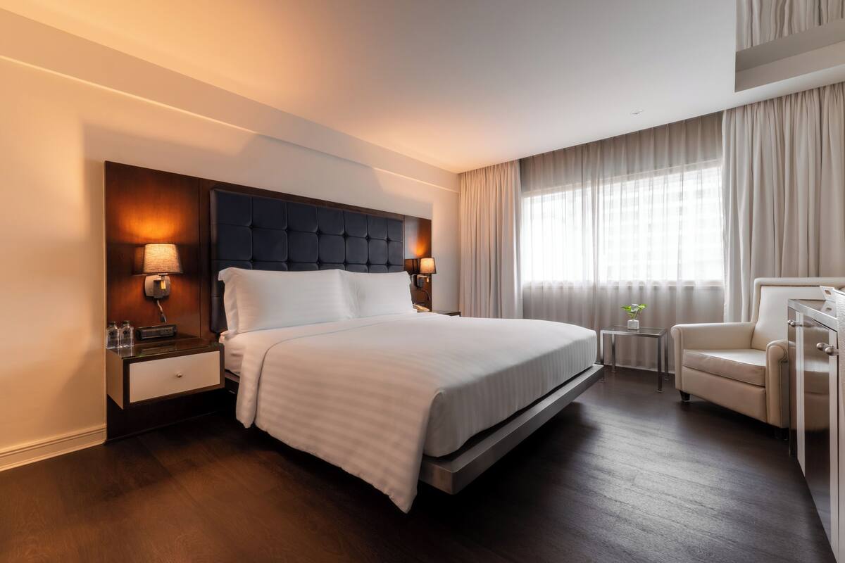 A modern hotel room with a large bed, bedside lamps, a small table, a chair, and a view of the city through the window.