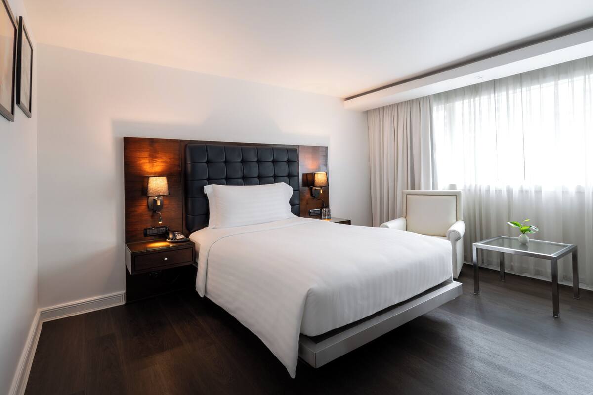 A modern hotel room with a large bed, a bedside table with a lamp, a chair, a small table, and a window with curtains.
