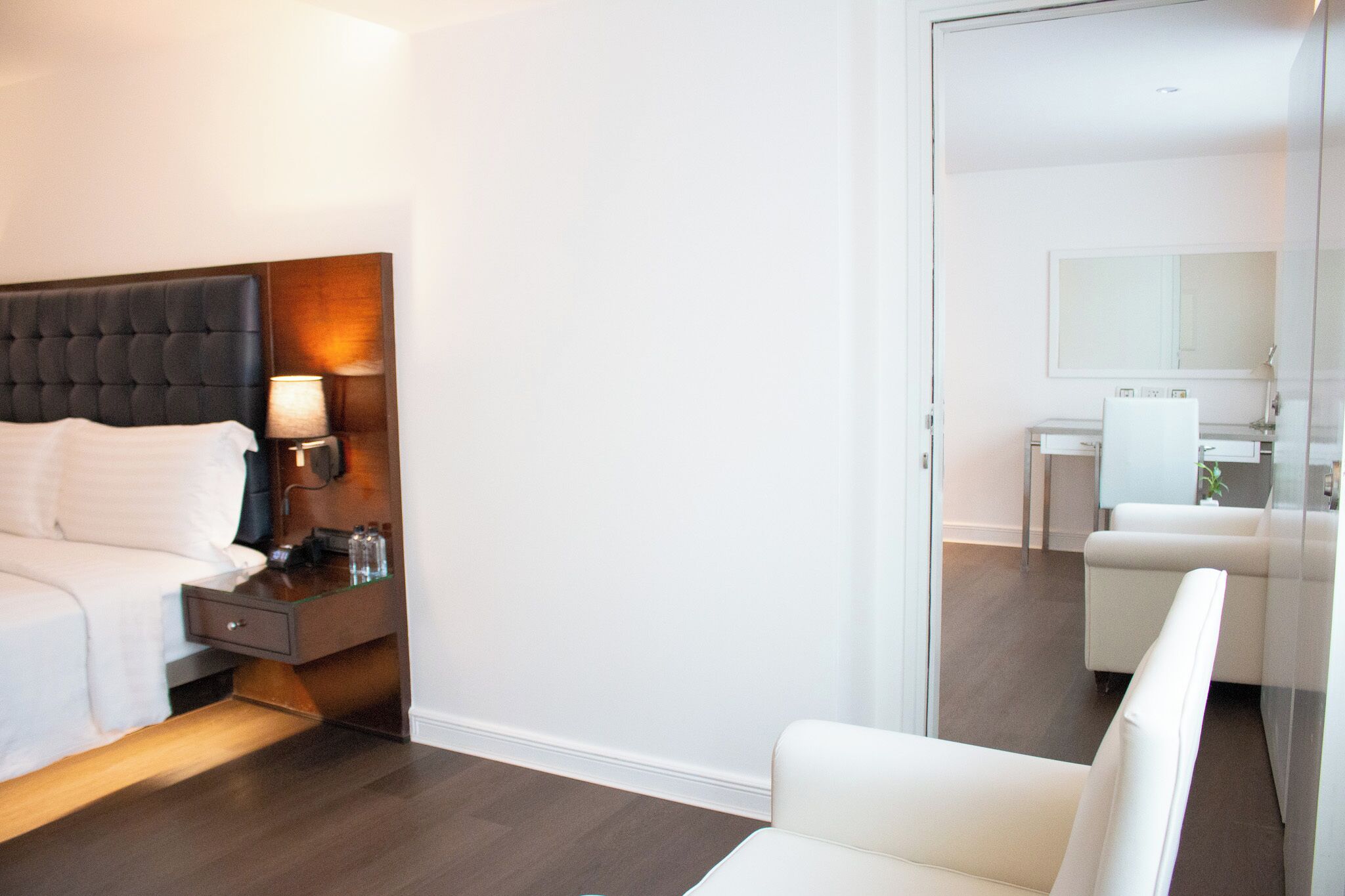 A hotel room with a bed, bedside table, lamp, and a view of a bathroom with a mirror and a chair.