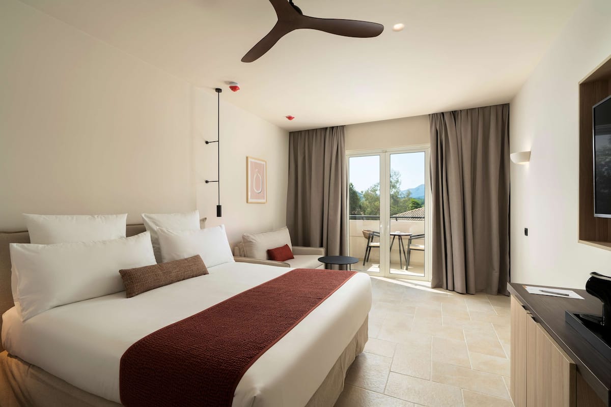 Habitación de hotel con una cama grande, un escritorio, una silla, un ventilador de techo y vistas al exterior a través de una puerta corrediza de vidrio.