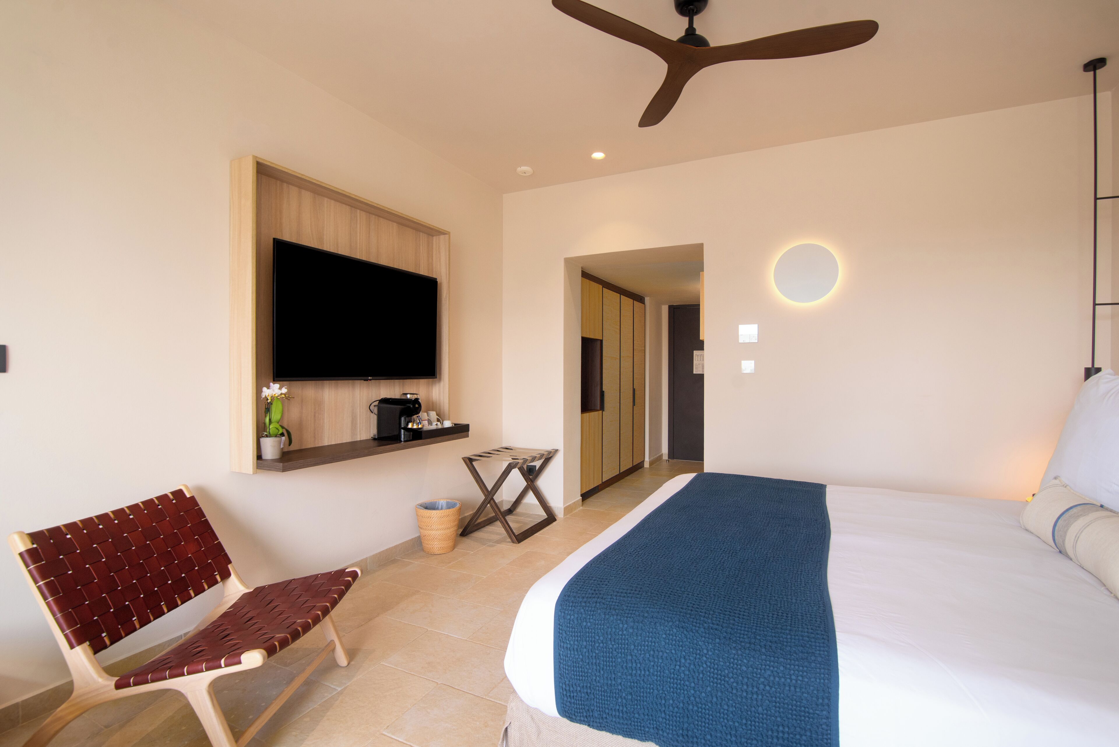 Habitación de hotel con cama, televisión instalada en la pared, ventilador de techo, una mesita con planta y un banco.