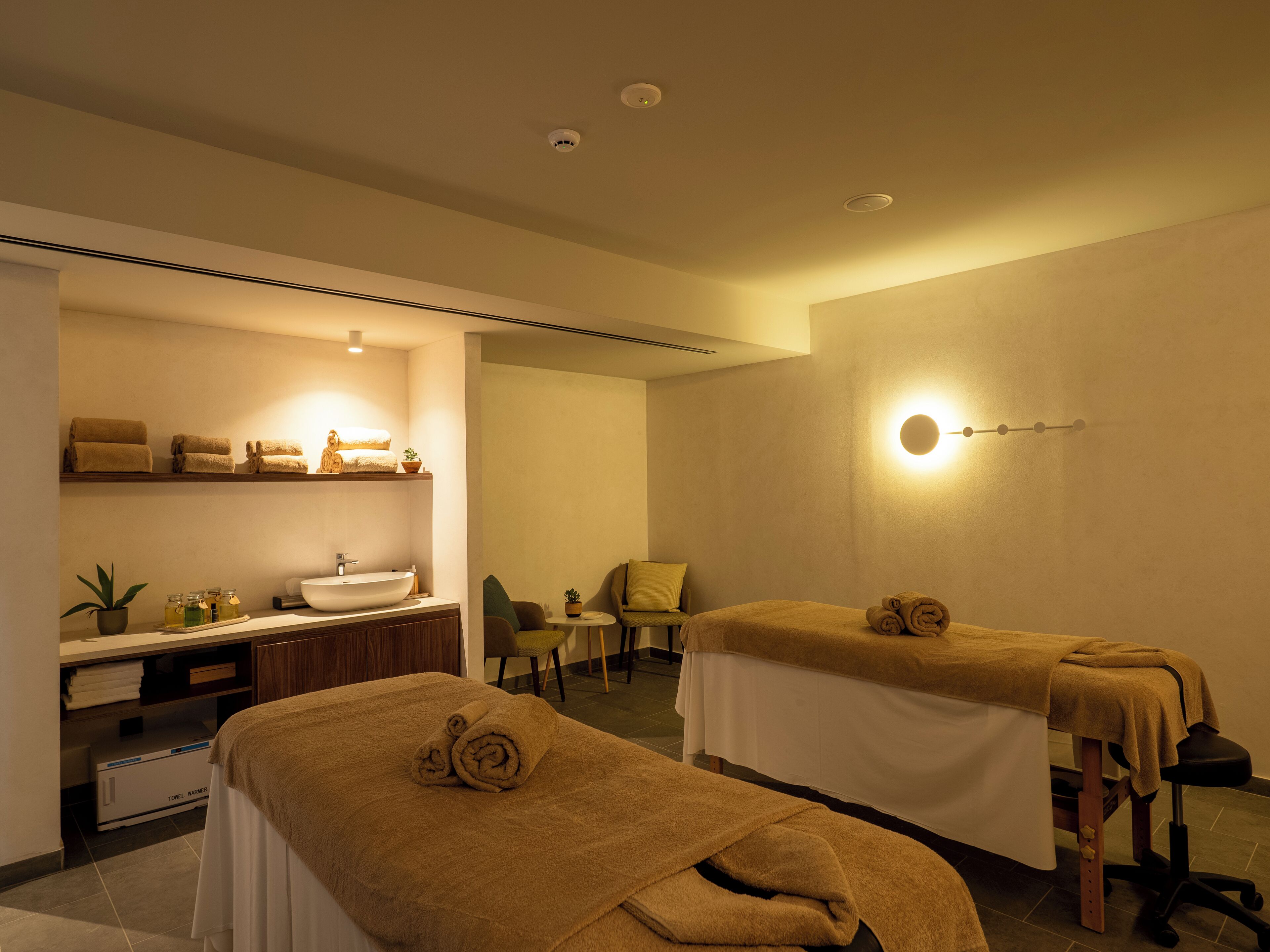 A spa room with two massage tables, a sink, and a shelf with towels.