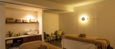 A spa room with two massage tables, a sink, and a shelf with towels.