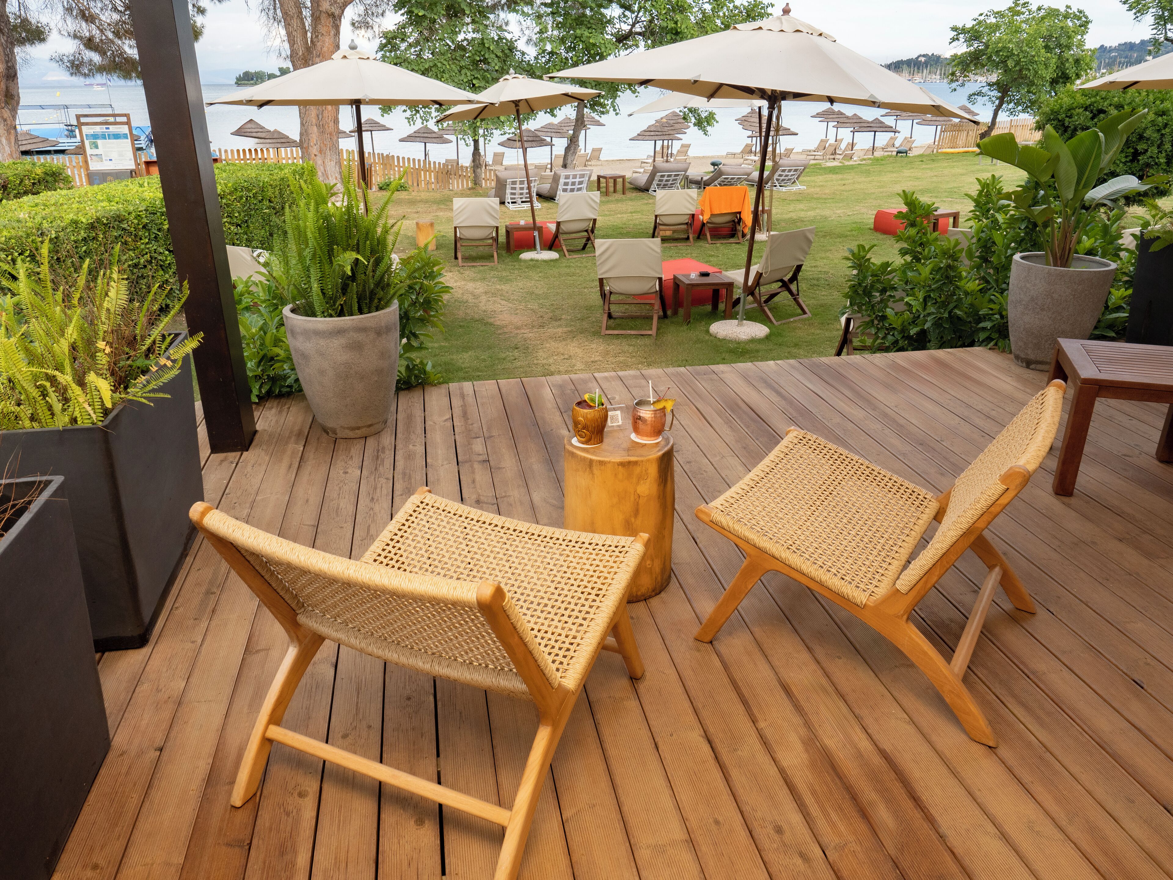 Una terraza de madera con sillas de mimbre y una pequeña mesa, con vistas a una zona de piscina con sombrillas y tumbonas.