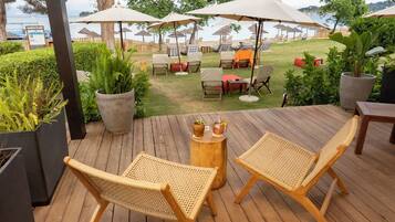 Una terraza de madera con sillas de mimbre y una pequeña mesa, con vistas a una zona de piscina con sombrillas y tumbonas.
