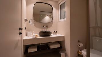 Et badeværelse med et rundt spejl, en vask med en stentop, toilet og skraldespand.