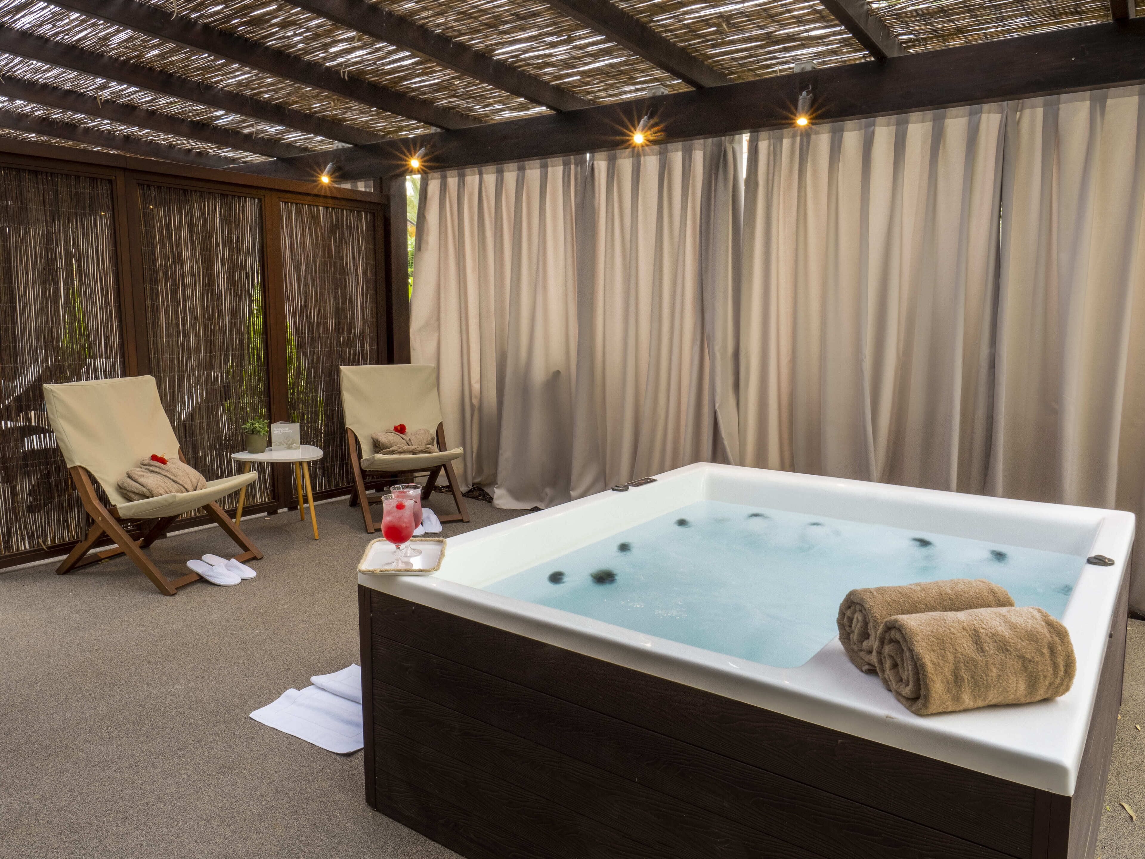 A spa area with a hot tub, two lounge chairs, a small table with a drink, and a curtain backdrop.