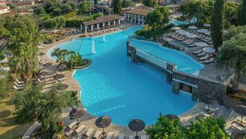 A large, curved swimming pool with a bridge, surrounded by lounge chairs and umbrellas, set in a lush, green environment.