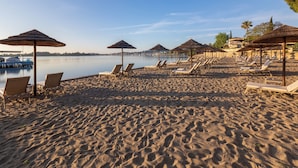 A beach with sun loungers and umbrellas, a calm lake, and a boat docked on the left.