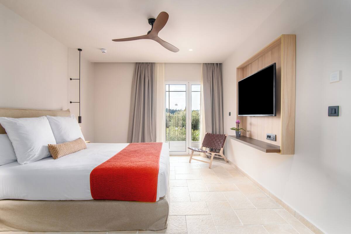 Una habitación de hotel moderna con una cama grande, un ventilador de techo, un televisor de pantalla plana montado en la pared y un ventanal corredero que da a un balcón.