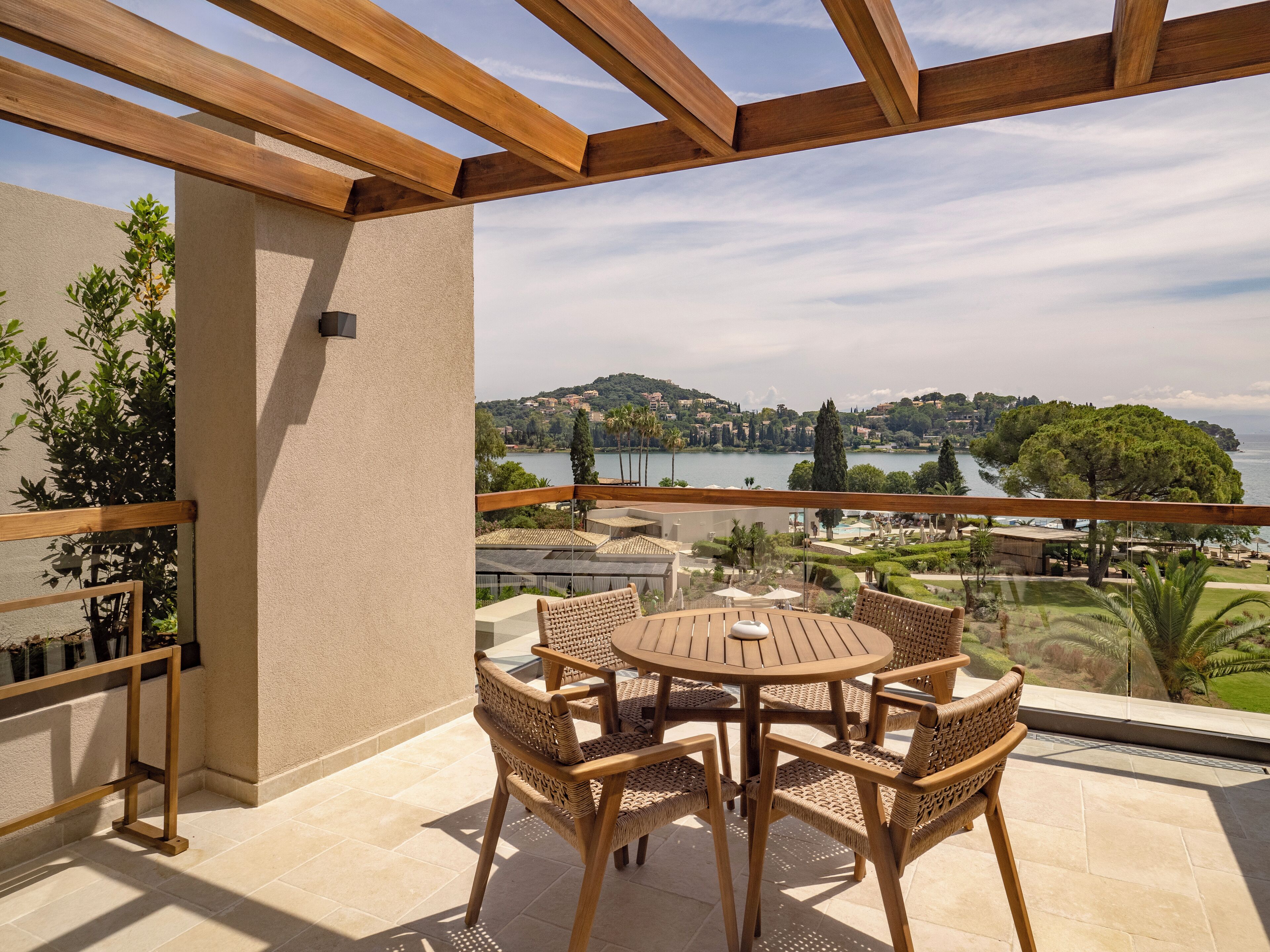 Un balcón con una pérgola de madera, mobiliario de mimbre y vistas a un lago y edificios a lo lejos.