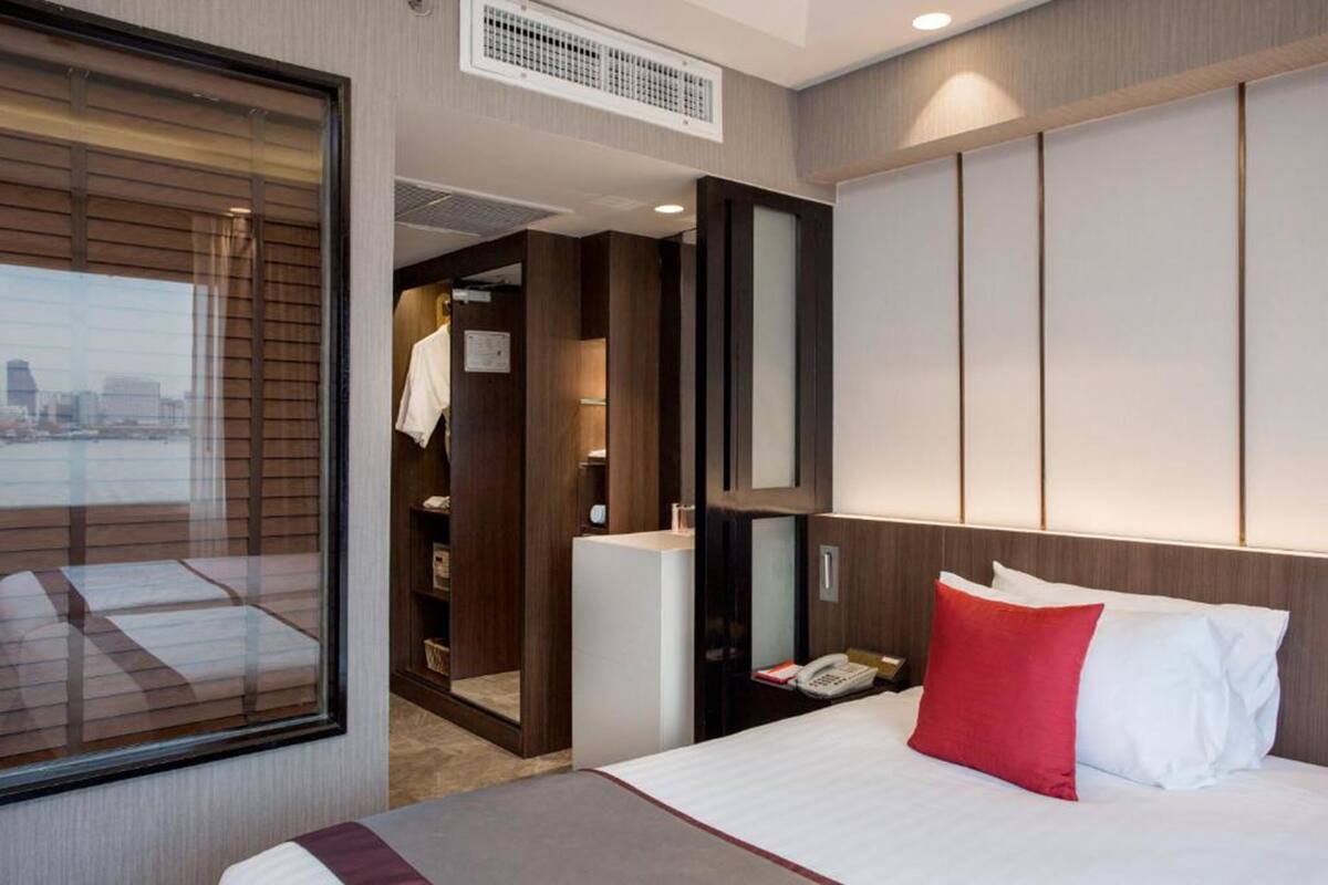 A hotel room with a large bed, a bedside telephone, a wardrobe, and a view of the city through a window.