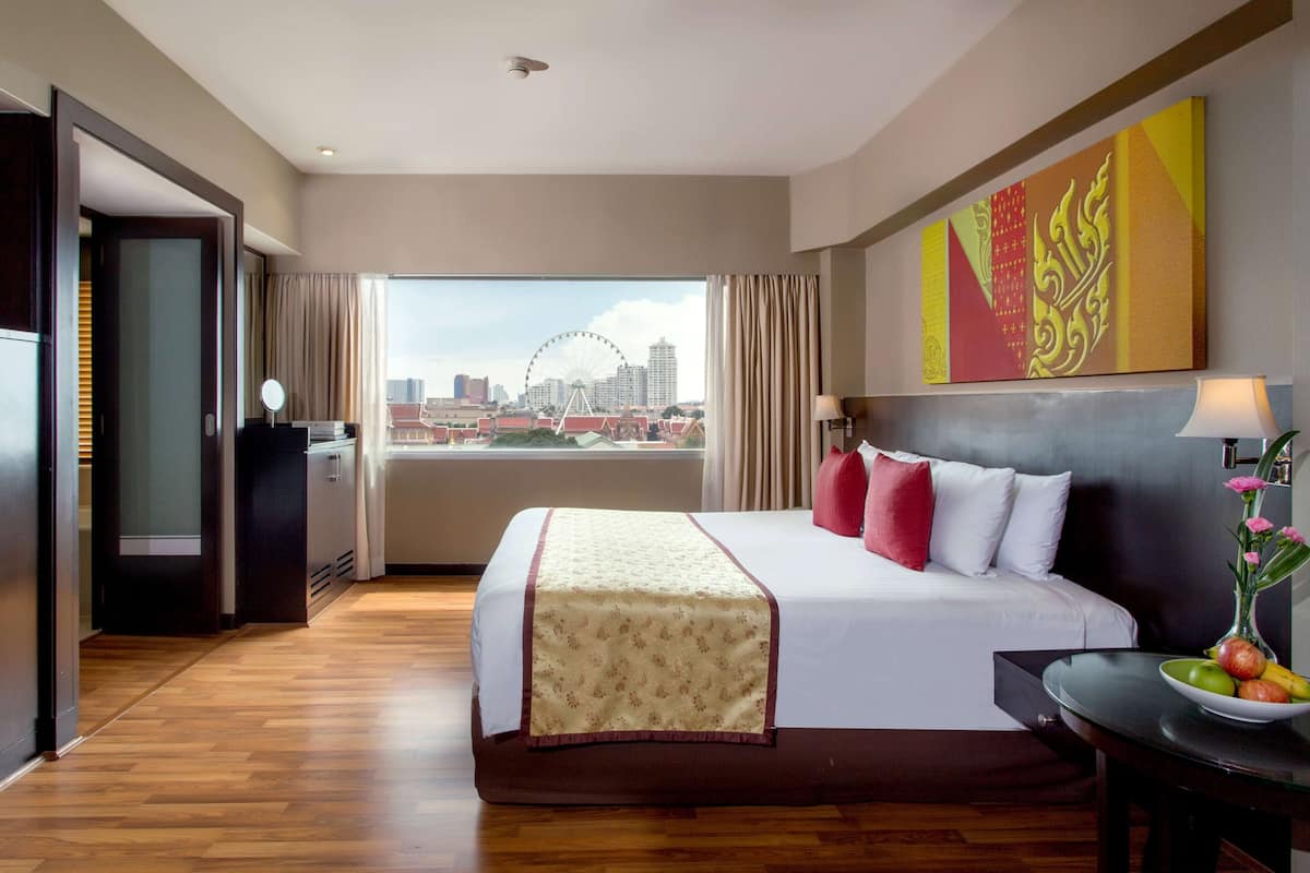 A hotel room with a large bed, a desk with a lamp, a view of a cityscape including a Ferris wheel, and a decorative wall piece.