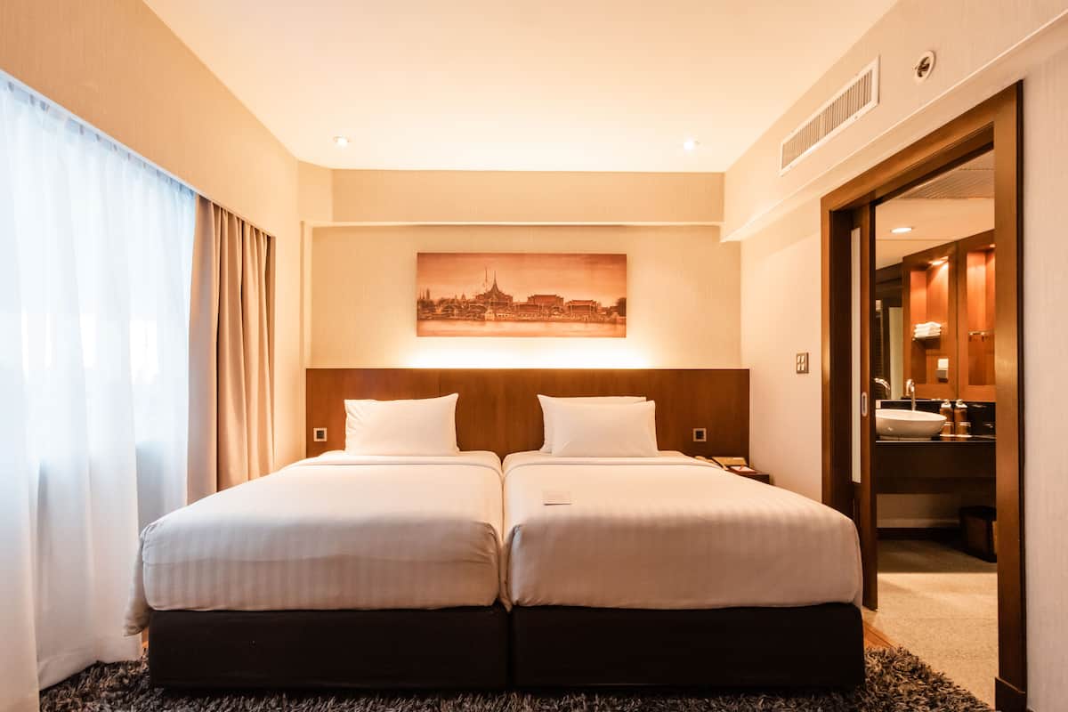 A hotel room with two beds, a cityscape painting on the wall, a bathroom visible through an open door, and a carpeted floor.