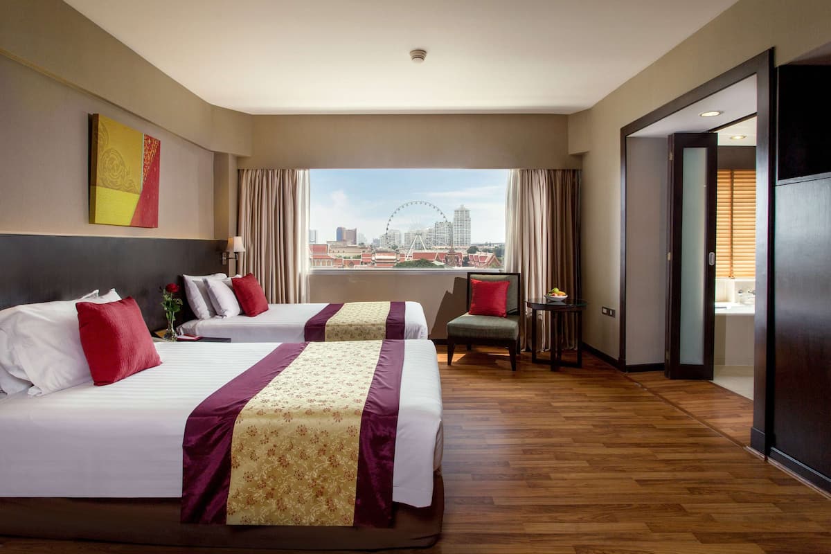 A hotel room with two beds, a view of a cityscape and a Ferris wheel, a small table with a lamp, and a chair.