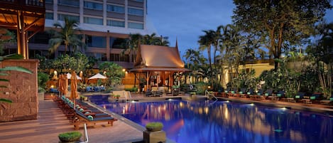 A swimming pool with lounge chairs and umbrellas, surrounded by palm trees and a building in the background.