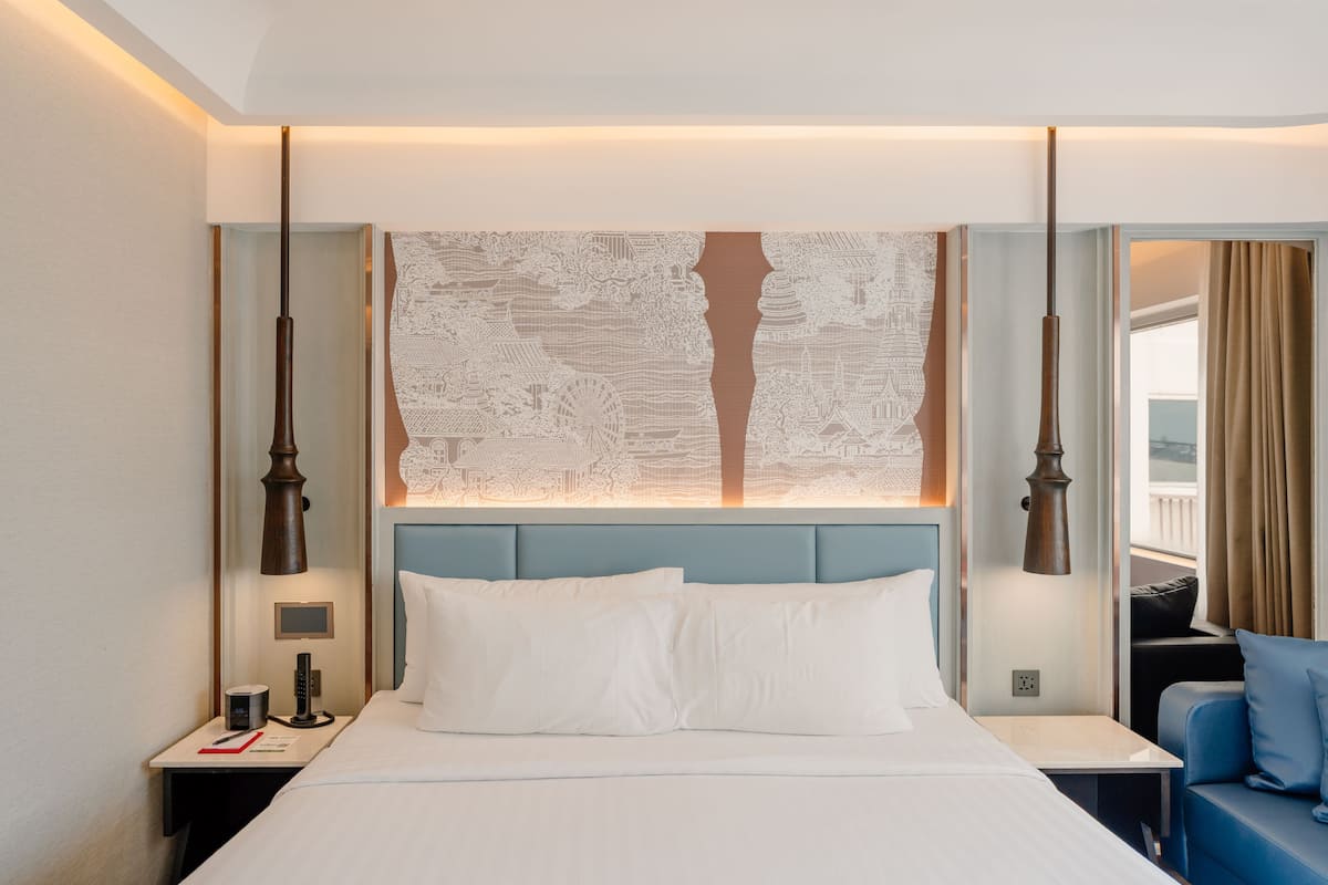 A hotel room with a large bed, a nightstand with a lamp, a small table with a cup and book, and a blue sofa.