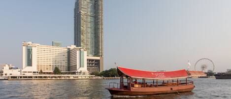 A river cruise boat with a red canopy, a Ferris wheel, and modern high-rise buildings in the background.