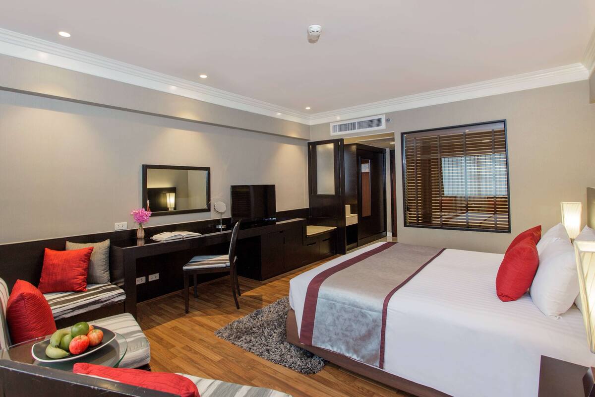 A hotel room with a bed, a desk, a chair, a sofa, a TV, and a window with blinds.