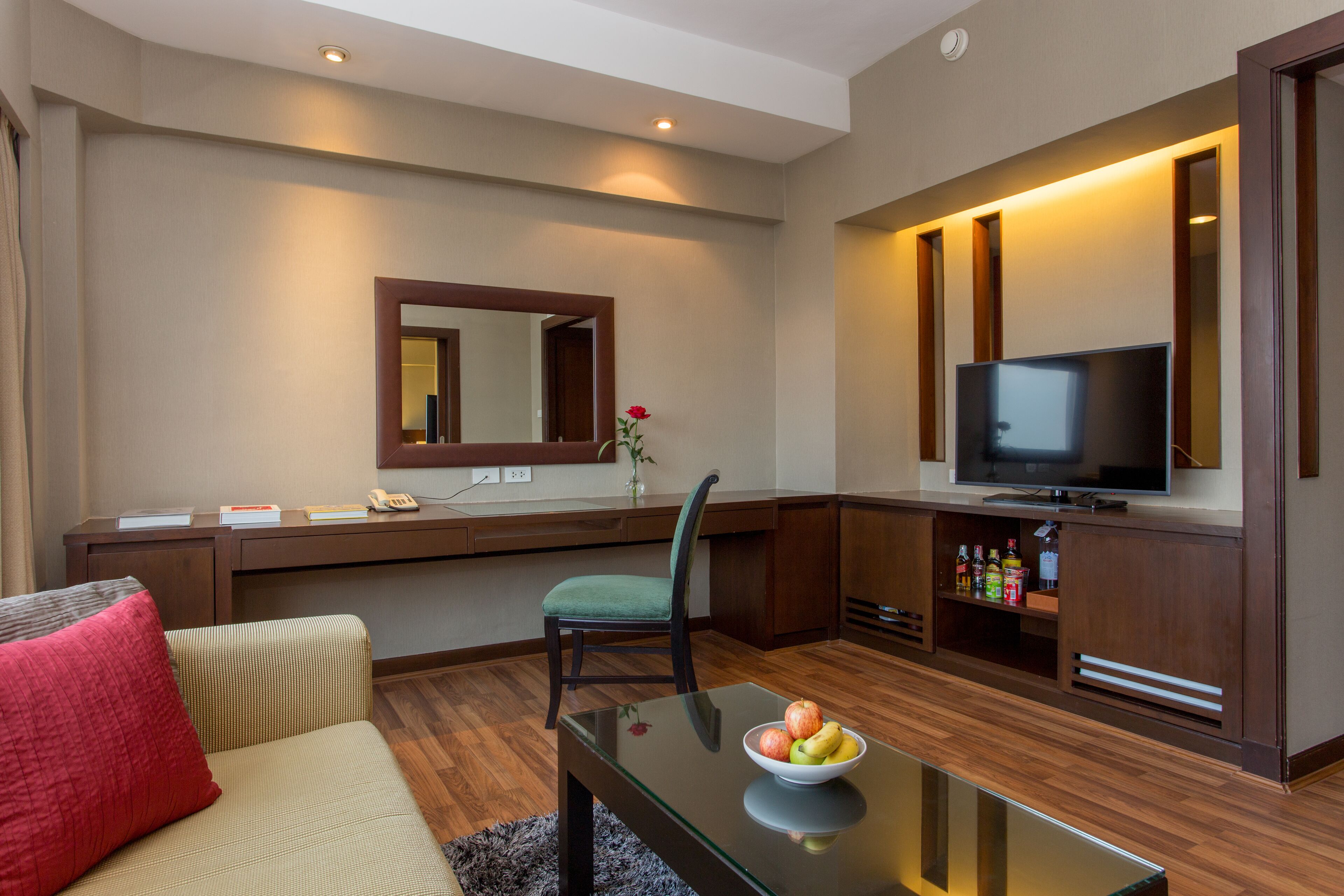 A modern hotel room with a sofa, a glass coffee table with fruit, a TV, a desk with a mirror, and a minibar.