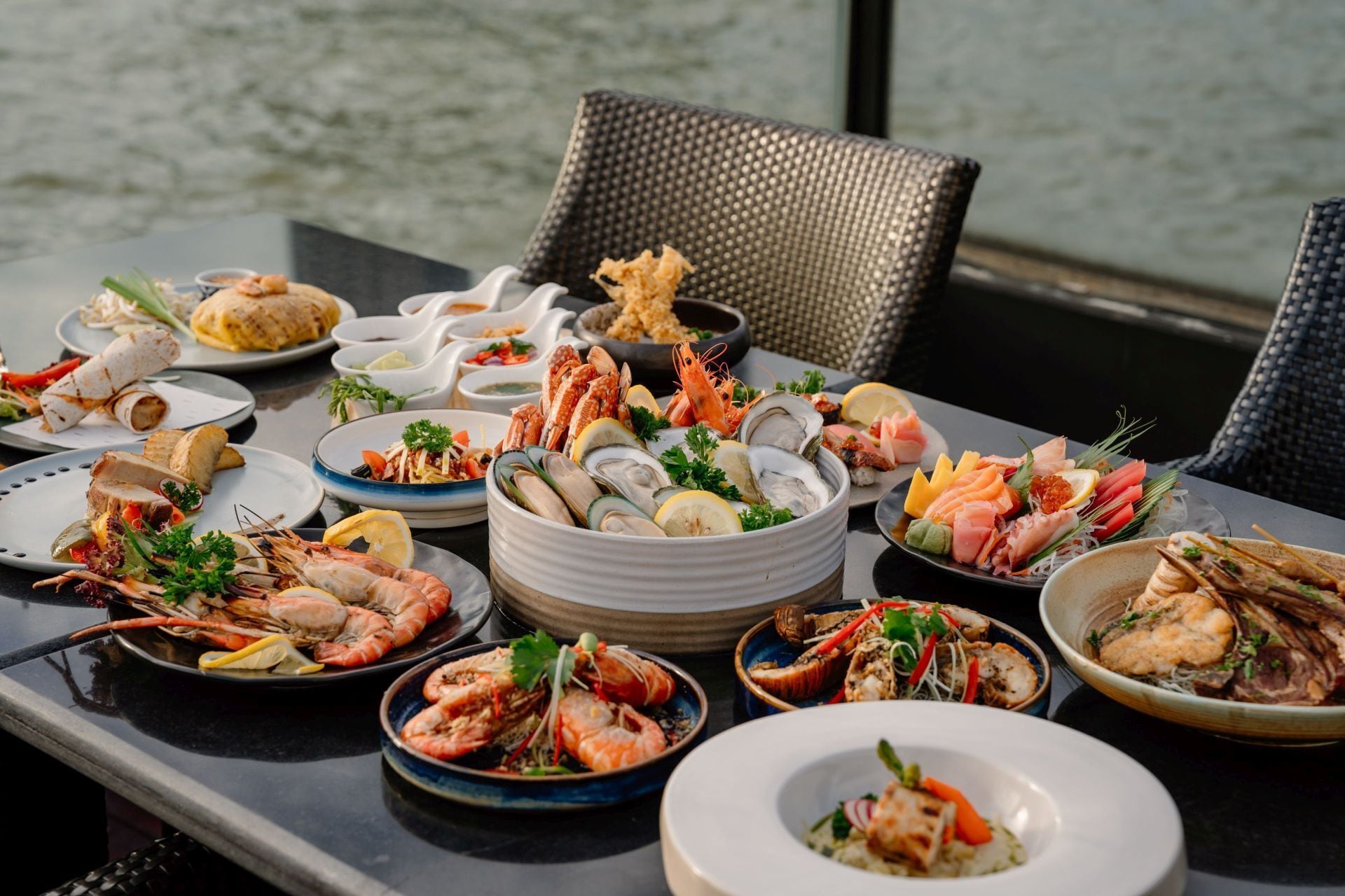Une table est dressée avec divers plats de fruits de mer, notamment des crevettes et des huîtres, et est placée de manière à offrir une vue sur l’eau en arrière-plan.