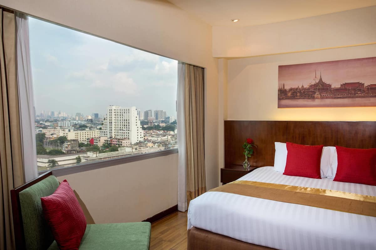 A hotel room with a large window, a bed with red and white bedding, a green chair, and a painting of a cityscape on the wall.