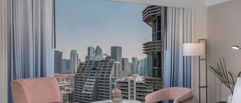 A hotel room with a city view, a bed, two chairs, a small table, and a floor lamp.