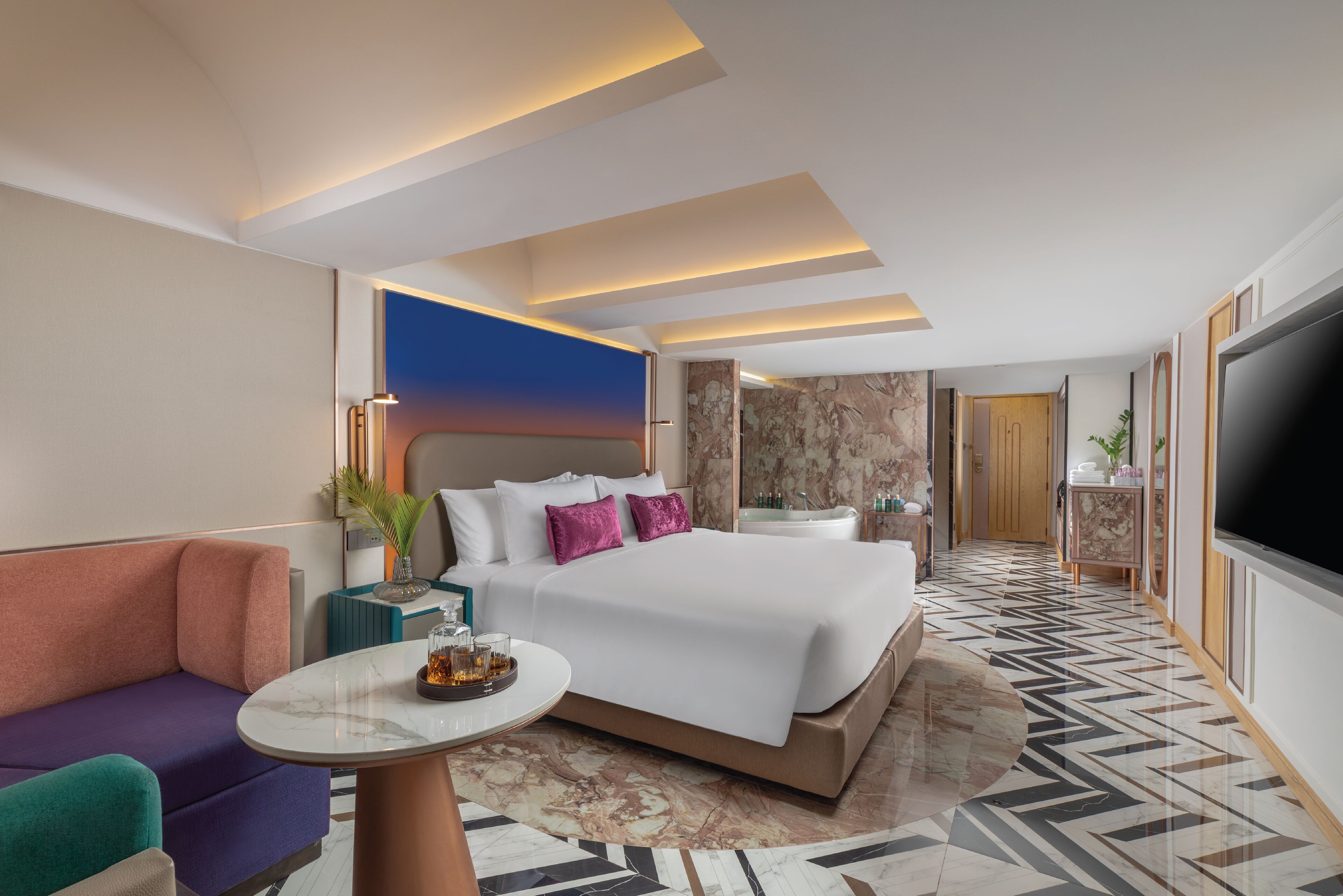 A modern hotel room with a large bed, a sofa, a TV, and a bathroom visible in the background.