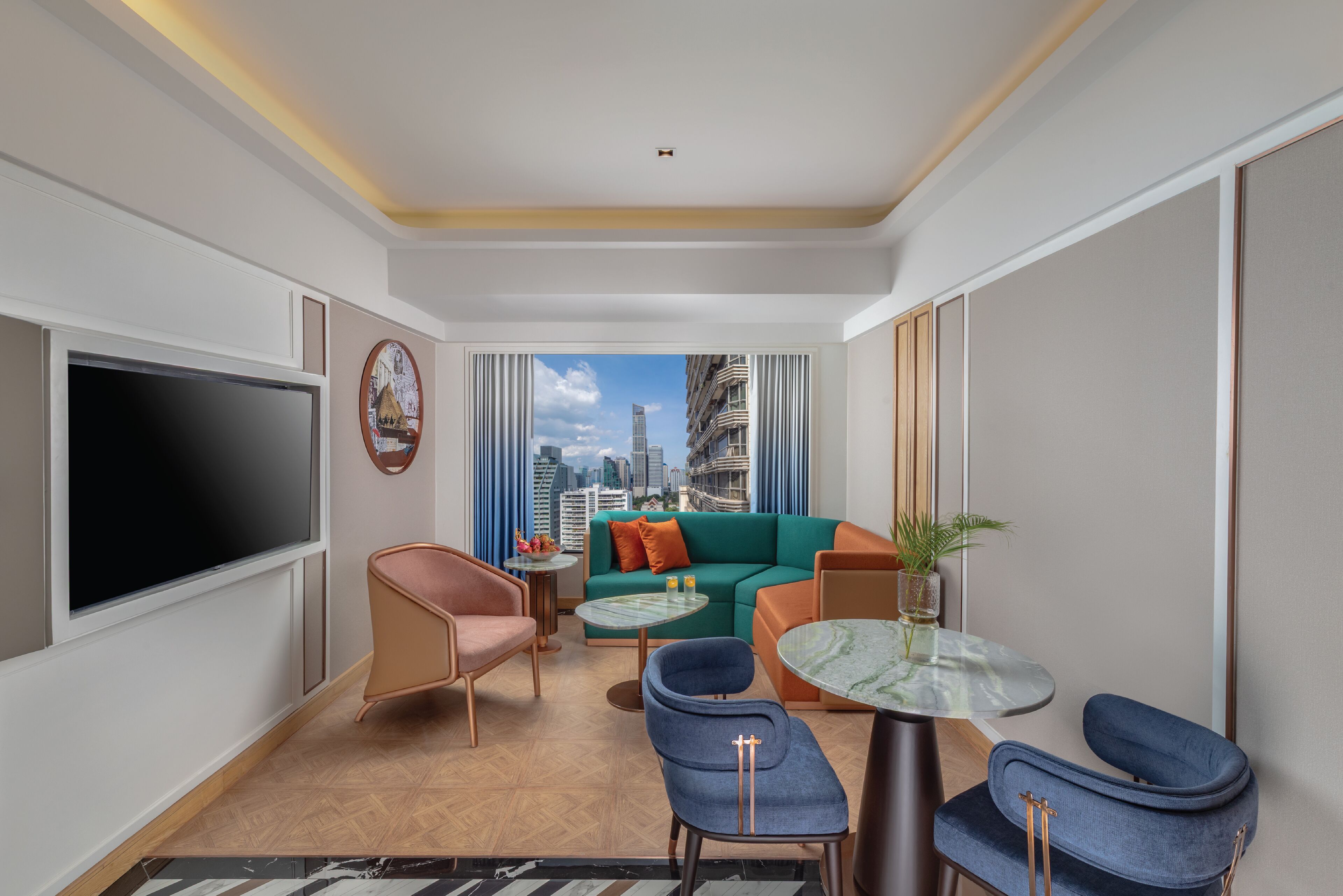 Ein modernes Hotelzimmer mit großem Fernseher, einem grünen Sofa, blauen Sesseln und Blick auf die Stadt.