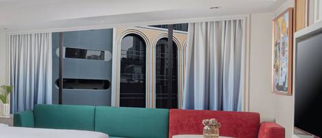 Una habitación de hotel moderna con un sofá verde aguamarina y rojo, una mesita redonda y un televisor en la pared derecha.