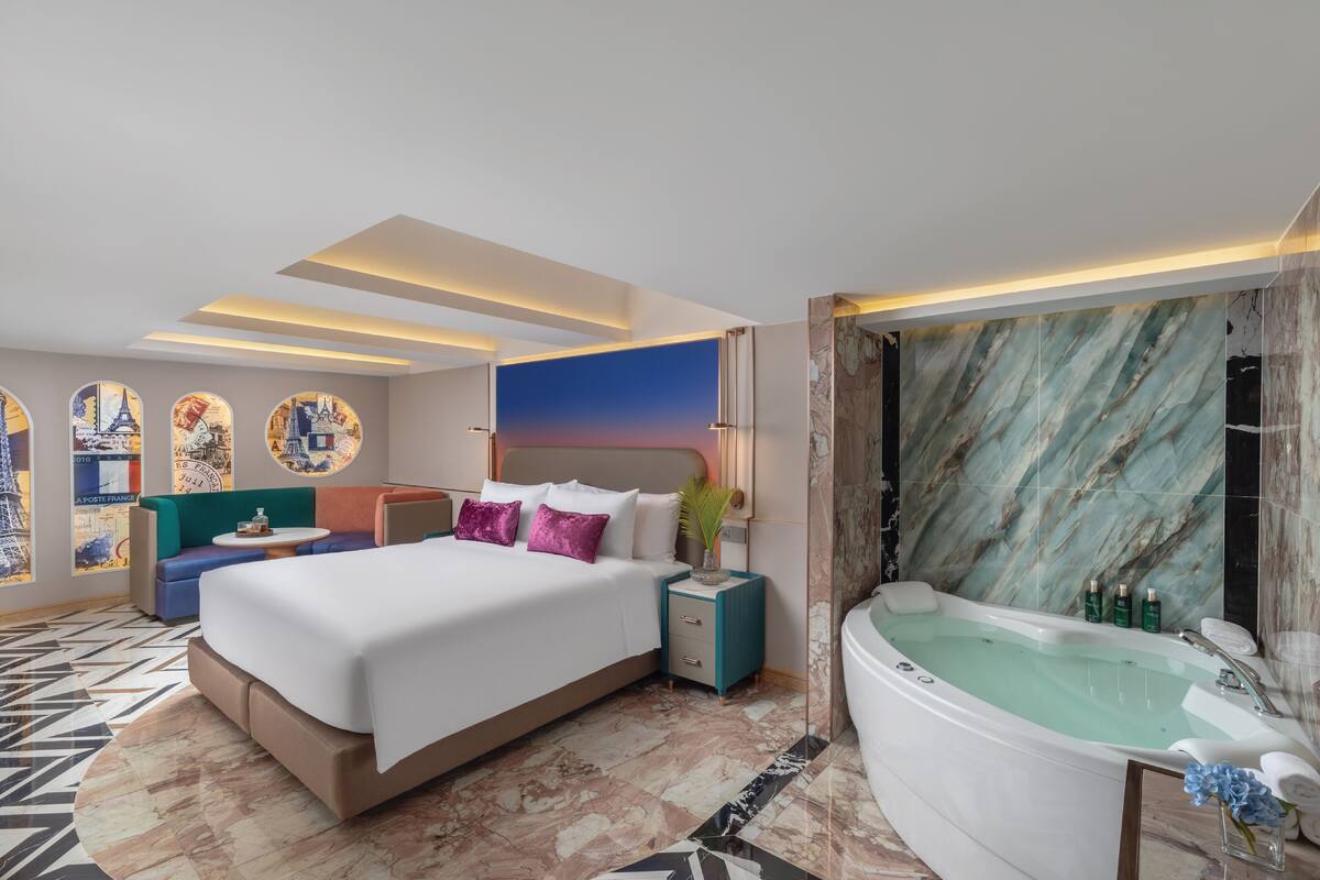 A modern hotel room with a large bed, a sitting area, a bathtub, and a cityscape mural on the wall.