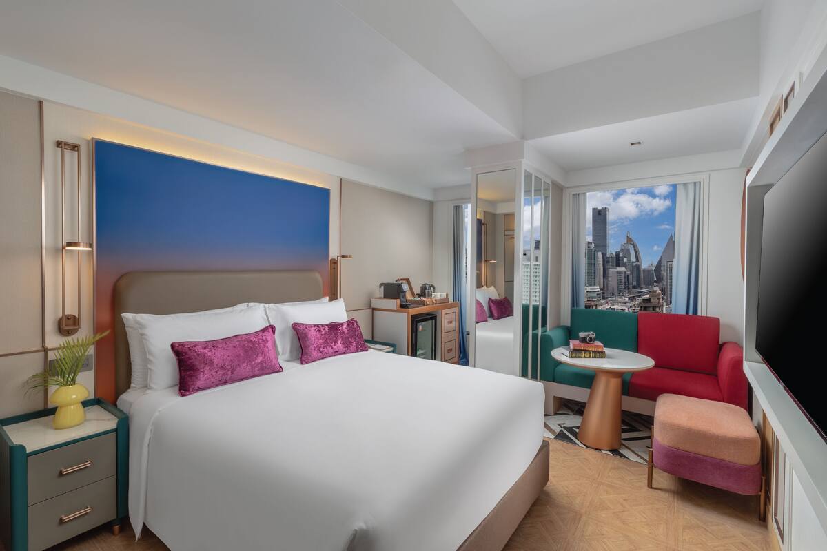 A modern hotel room with a large bed, a desk, a red sofa, a TV, and a cityscape view.