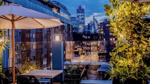 An outdoor seating area with tables and chairs, a large umbrella, and a view of a cityscape with modern buildings. - Pipa Bangkok Sukhumvit 11 by Kingston Hotels (Bangkok)