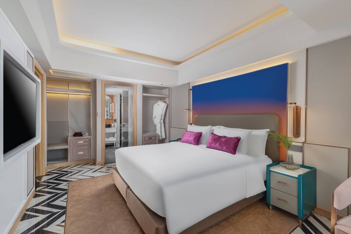 A modern hotel room with a large bed, a TV, a walk-in closet, and a painting on the wall.