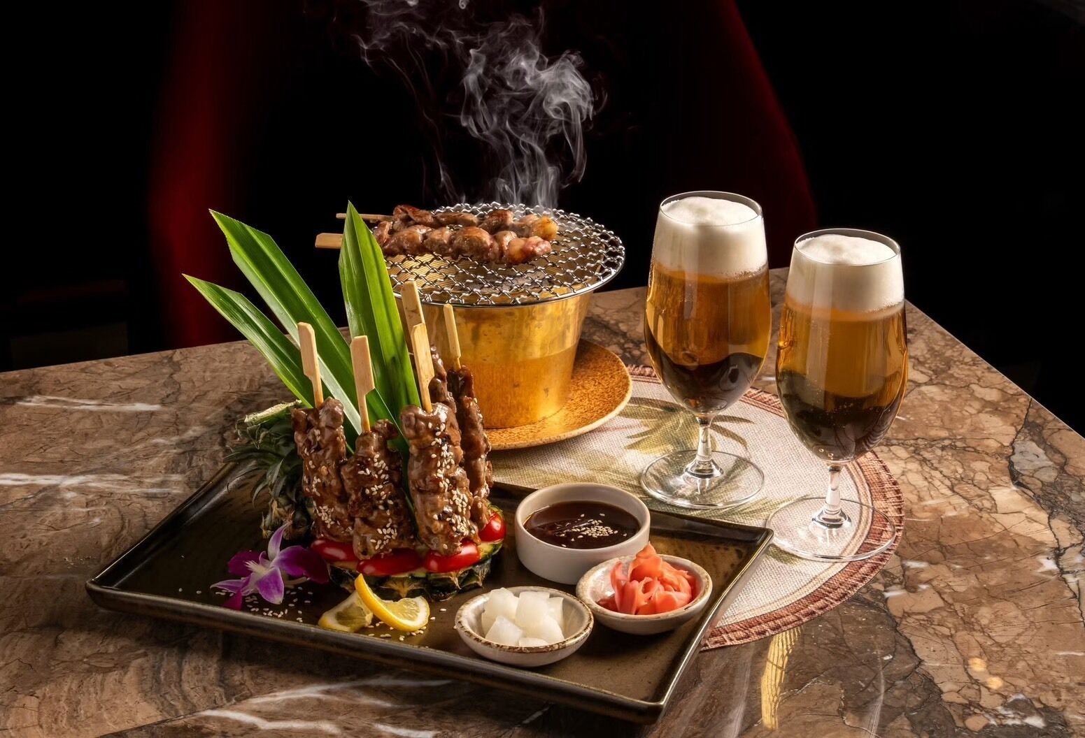 Una bandeja con brochetas, un recipiente con salsa y dos vasos de cerveza sobre una mesa de mármol.